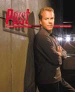 A man with blond hair and a gray shirt leans against a wall. A red logo is visible behind him.