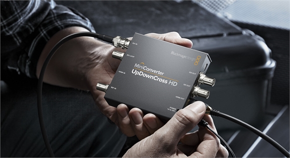 A person holds a small black Blackmagic Design MiniConverter device with several SDI and HDMI connections.