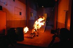 A large fire burns on a wooden pallet in a dark, industrial-style room.
