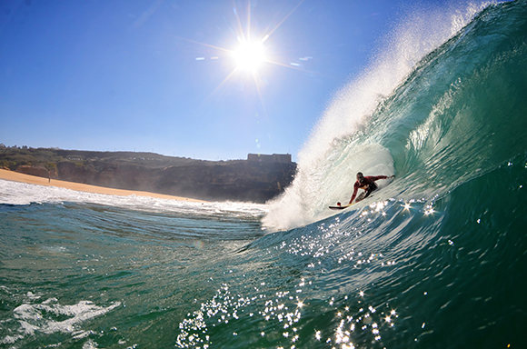 A surfer rides inside the barrel of a large, turquoise ocean wave under a bright, sunny sky.