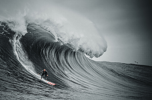 A surfer in a black wetsuit rides a giant, curling ocean wave under a cloudy sky.