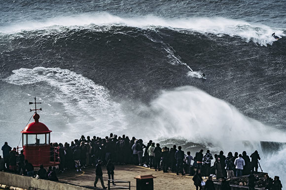 A giant dark wave crests and breaks near a red lighthouse structure and a crowd of people on a pier.