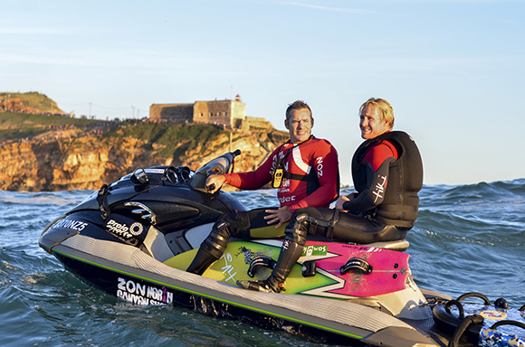 Two men in wetsuits riding a black and yellow jet ski on the ocean near a cliffside fort.