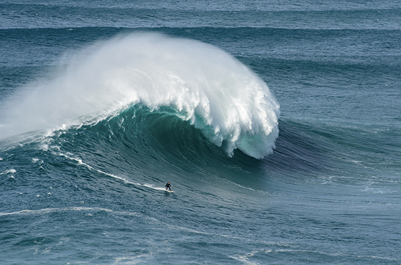 A lone surfer rides the face of a giant, curling turquoise ocean wave with white spray.
