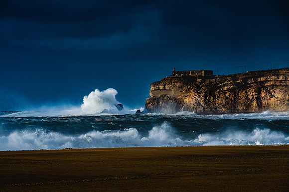 Large white waves crashing against a steep rocky cliff under a dark, stormy sky.