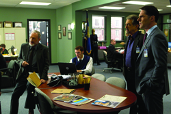 Three men in professional attire stand in a green-walled office around a conference table.