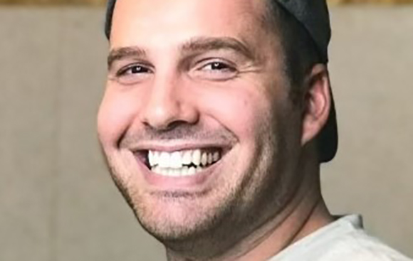 A man with short dark hair and stubble smiling broadly while wearing a dark baseball cap.