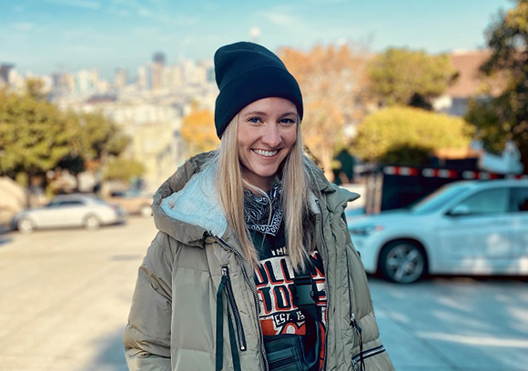 A smiling blonde woman wearing a black beanie and an olive green puffer jacket in an urban setting.