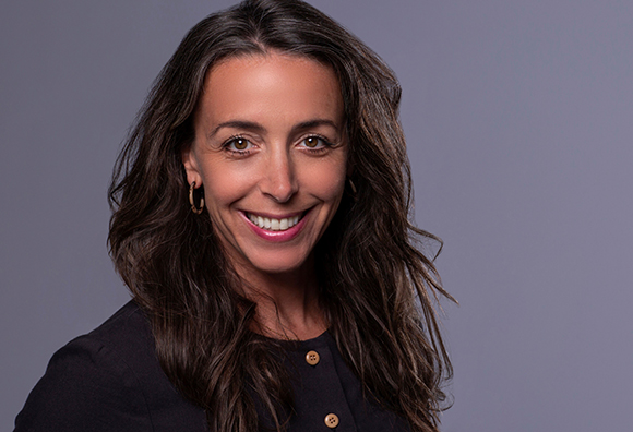 A smiling woman with long wavy brown hair and gold hoop earrings wearing a dark button-up shirt.