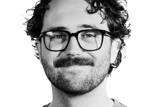 Black and white close-up portrait of a smiling man with curly hair, glasses, and a mustache.