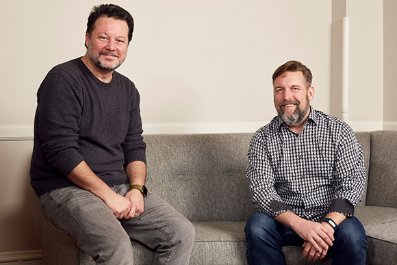 Two men with facial hair sit on a gray couch against a plain white wall.