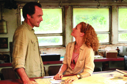 A man in a tan shirt and a woman with curly red hair look at each other over a table near windows.