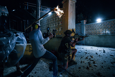 Film crew filming a night scene with a man kneeling and holding a prop weapon near a flare.