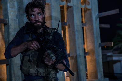A bearded man in tactical gear holds a rifle while looking off to the side in a dimly lit structure.