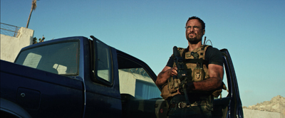 A man in a tan tactical vest holding a rifle stands next to an open dark blue pickup truck.