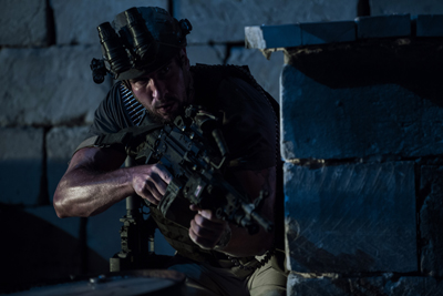 A soldier wearing night vision goggles aims a rifle from behind a stone wall in low light.