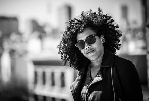 Black and white portrait of a person with voluminous curly hair and sunglasses, tilting their head.