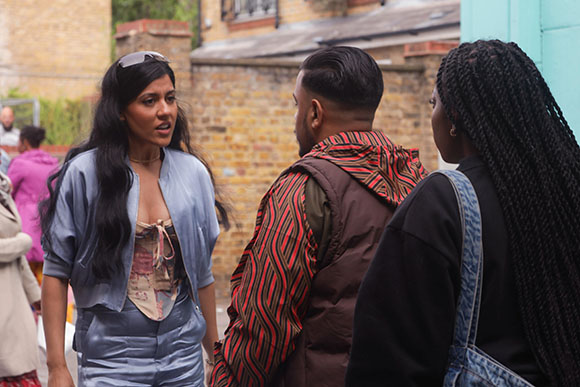 A woman in a blue satin outfit speaks to a man in a patterned jacket and a woman with long braids.