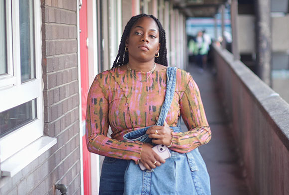 A woman with braided hair wearing a colorful patterned top and denim bag stands on an outdoor walkway.