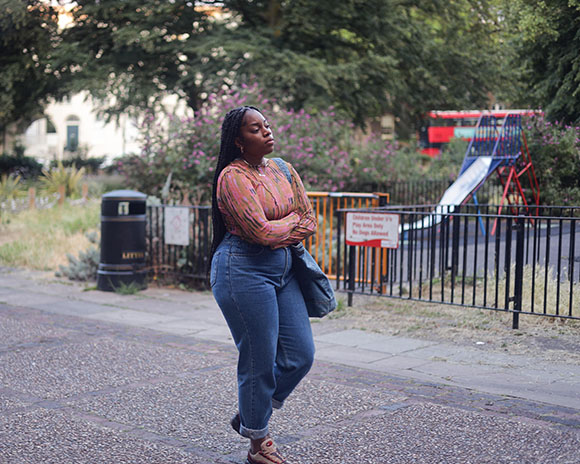 A woman with long braids and a patterned shirt stands with eyes closed in a park near a playground.