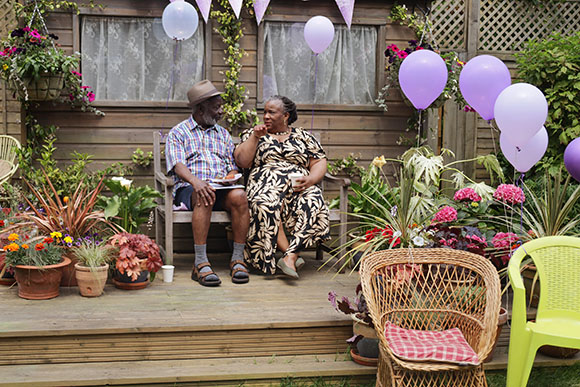 An older man and woman sit on a garden bench surrounded by flowers and purple balloons.