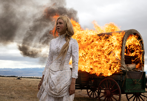 A blonde woman in a white period dress stands before a wooden wagon engulfed in large orange flames.