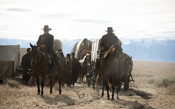 People on horseback and in covered wagons travel across a vast, dry plain toward distant mountains.