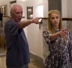 An older man in a blue shirt and a blonde woman in a plaid dress both point their fingers forward.