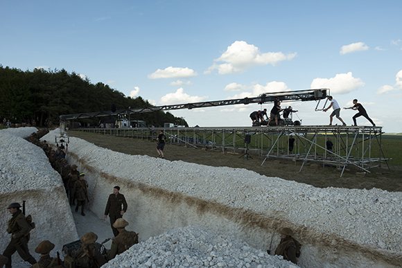 A film crew operates a long camera crane over a trench set with actors in period military uniforms.