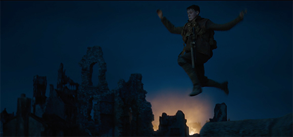 A person in military attire jumps mid-air with arms outstretched against a dark night sky and stone ruins.