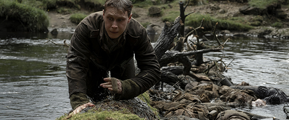 A man in a dark jacket crawls from a river onto a muddy bank filled with debris.