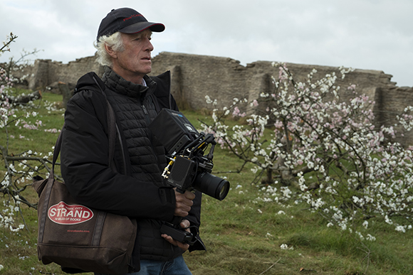 A man in a black jacket and cap holds a professional video camera in a grassy field with white blossoms.