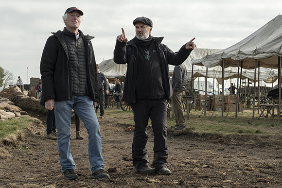 Two men in jackets and hats stand on a dirt path near large white canvas tents.
