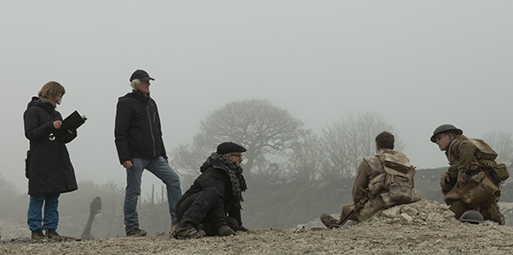 A film crew stands and sits on a dirt mound in heavy fog near actors in military gear.
