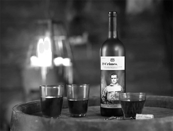 A bottle of 19 Crimes wine stands next to three filled glasses on a wooden barrel in black and white.