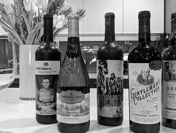 Five different wine bottles standing in a row on a white countertop next to a vase of flowers.