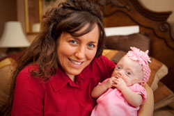 A smiling woman in a red shirt holds a baby wearing a pink headband and outfit.