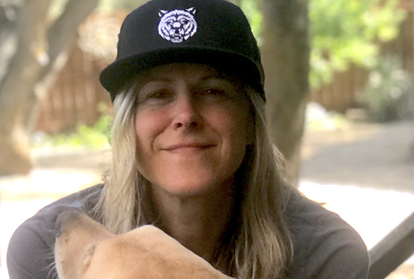 A smiling woman with blonde hair wearing a black baseball cap with a white animal logo.