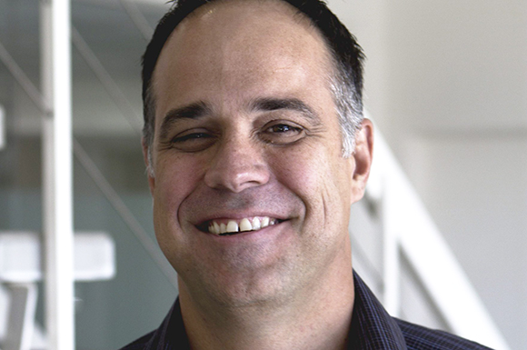 A close-up portrait of a middle-aged man with short dark hair smiling.