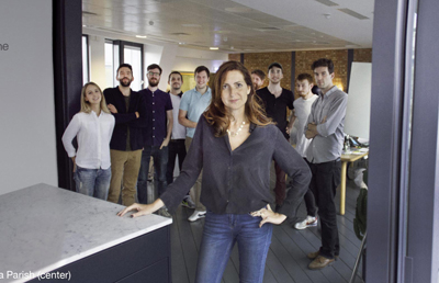 A woman in a dark shirt and jeans stands in the foreground of a group of people in an office.