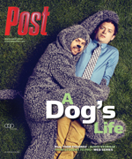 A man in a fuzzy dog costume embraces another man wearing a shirt and tie, both lying on green grass.