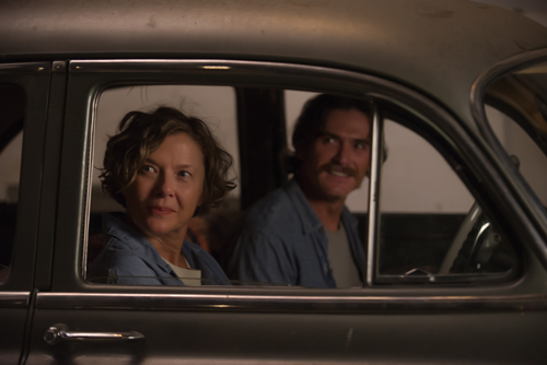A woman with short wavy hair and a man with a mustache look out from the window of a vintage car.