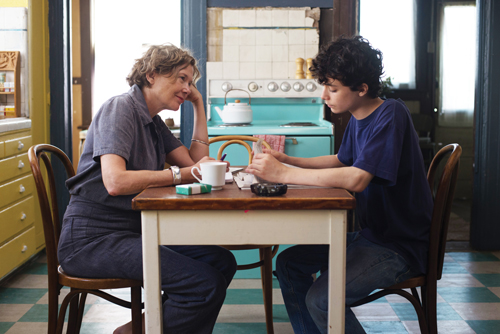 A woman and a young person sit at a small wooden table in a kitchen, talking.