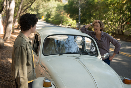 A young person in a green jacket looks at a woman leaning against a white vintage Volkswagen Beetle.