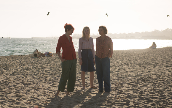Three people stand on a sandy beach facing the ocean under a bright sky with birds flying above.