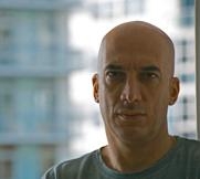 A close-up portrait of a bald man with a serious expression against a blurred urban background.
