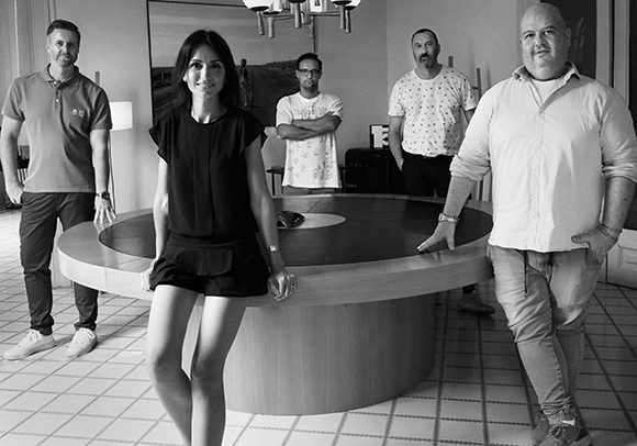 Five people stand around a large round table in a modern indoor setting, captured in black and white.