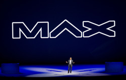A man in a suit stands on a stage with blue lighting. A large white "MAX" logo is displayed on a dark blue background.