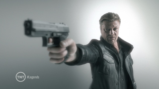 A man in a dark leather jacket points a handgun directly at the camera against a gray background.