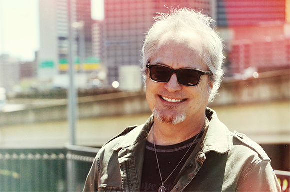 A smiling man with grey hair and dark sunglasses wearing a green vest.